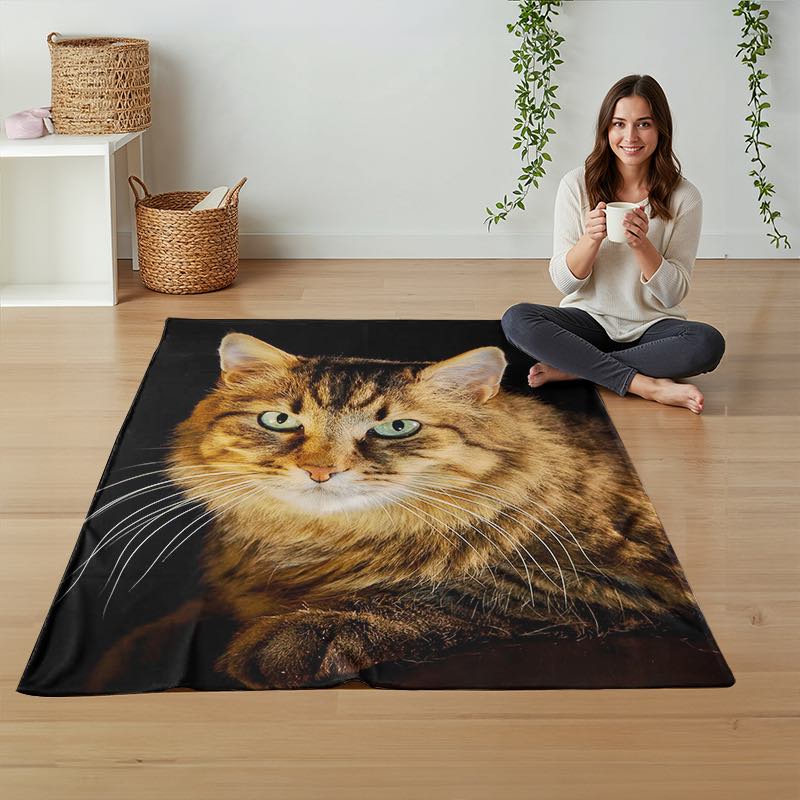A woman sits cross-legged on a light wood floor next to a large rectangular throw blanket featuring a realistic printed image of a fluffy brown tabby cat with green eyes. The scene is styled with woven baskets and hanging greenery against a white wall.