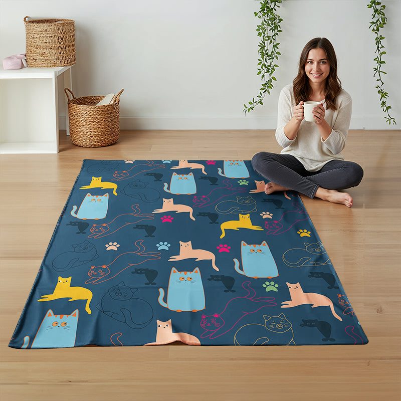 A woman sits cross-legged on a light wood floor next to a large navy blue fleece blanket printed with colorful cartoon cats and paw prints. The room has a white wall, a small white side table with woven baskets, and hanging green vines.