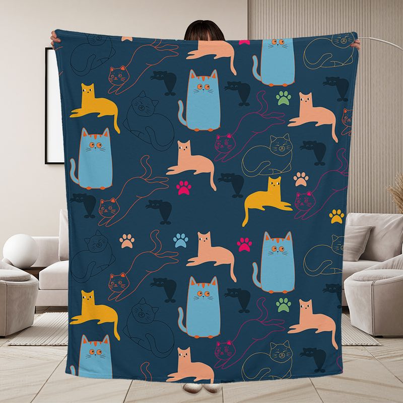 A large throw blanket with a dark navy blue background with a playful repeating pattern of cartoon cats in light blue, peach, yellow, and black, along with paw prints, held by a person in the living room setting.