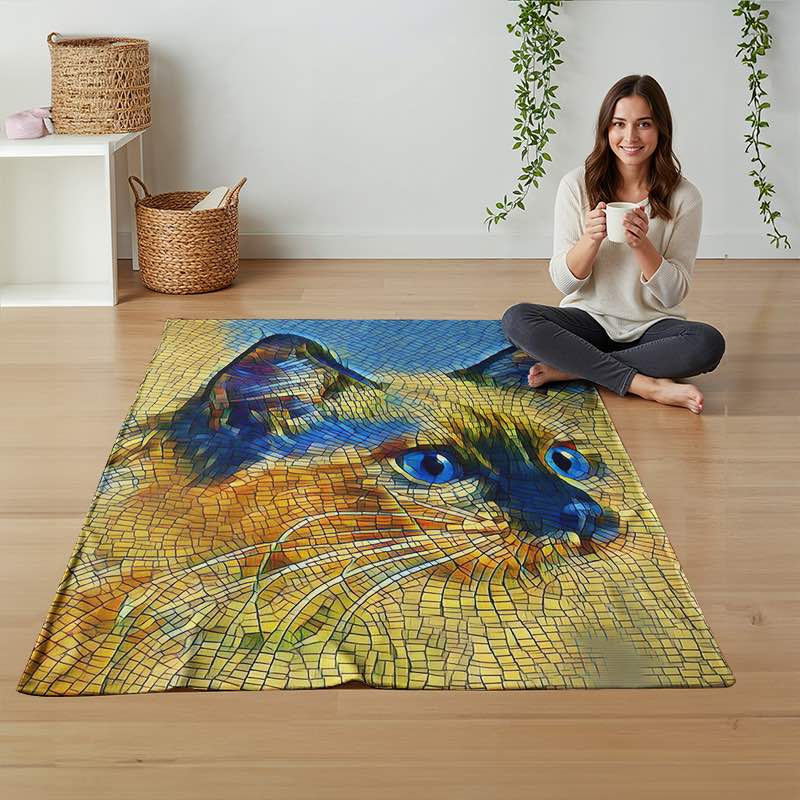 A woman sits cross-legged on a light wood floor next to a large rectangular throw blanket featuring a mosaic-style cat face with vivid blue eyes and yellow, orange, and blue tones. The room has a white wall, hanging greenery, woven baskets, and a small white side table.