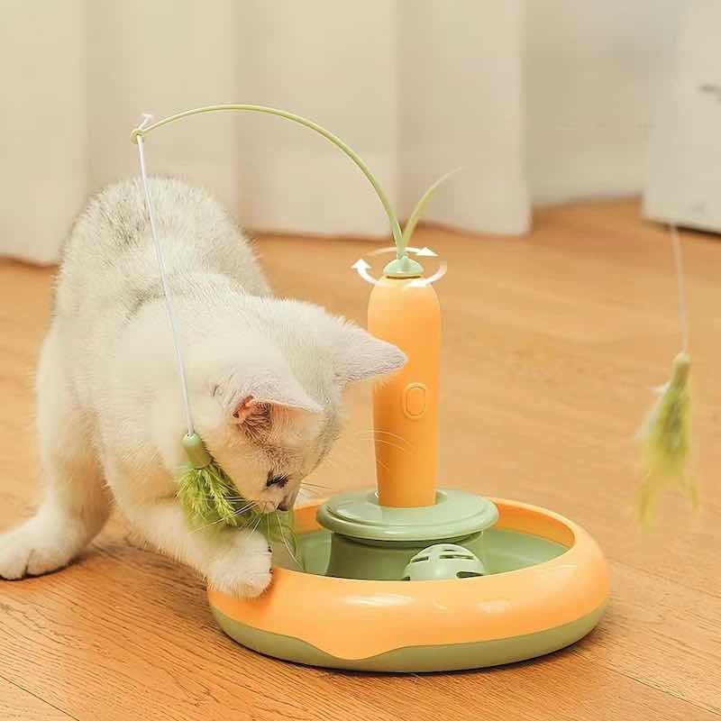 A white kitten playing with an orange and green multi-functional cat toy on a wooden floor.